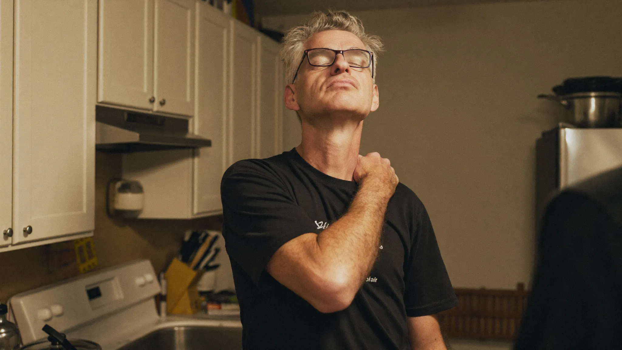 A man standing alone in his kitchen in the evening, hand on the back of his neck